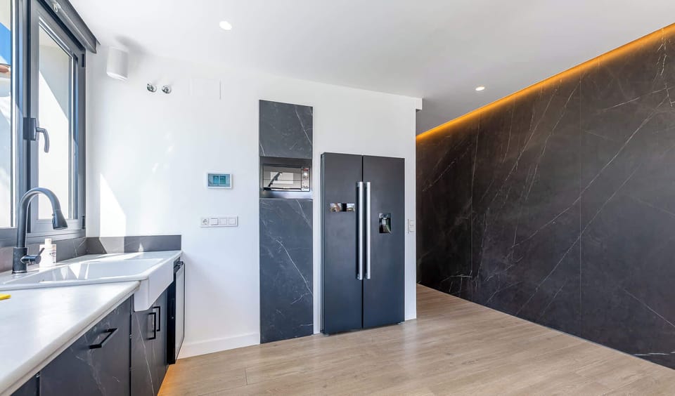 Sleek kitchen with black-paneled fridge, veined tile walls, and warm wood flooring. Natural light and ambient LED accents highlight the modern design, blending elegance with everyday functionality.