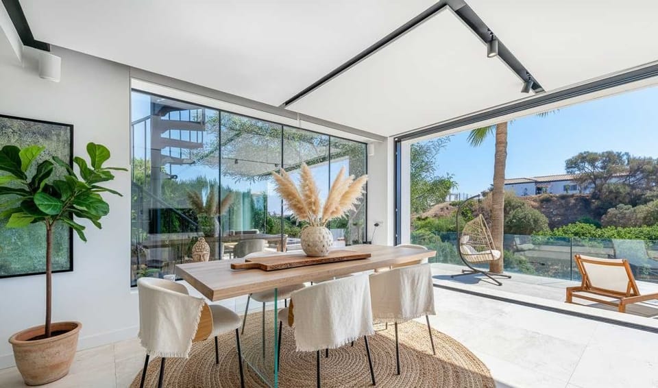A sunlit dining space with floor-to-ceiling windows, natural wood table, white chairs, and lush garden views. A woven rug and pampas centerpiece add warmth, blending indoor elegance with outdoor serenity.