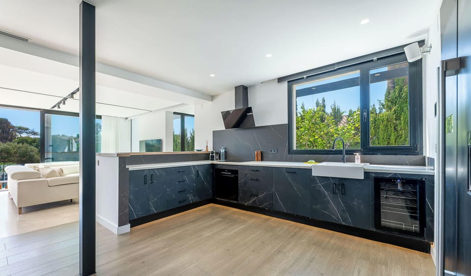 Modern kitchen with black marble counters, white farmhouse sink, and light wood floors. Large windows brighten the space, while clean lines and dark cabinetry