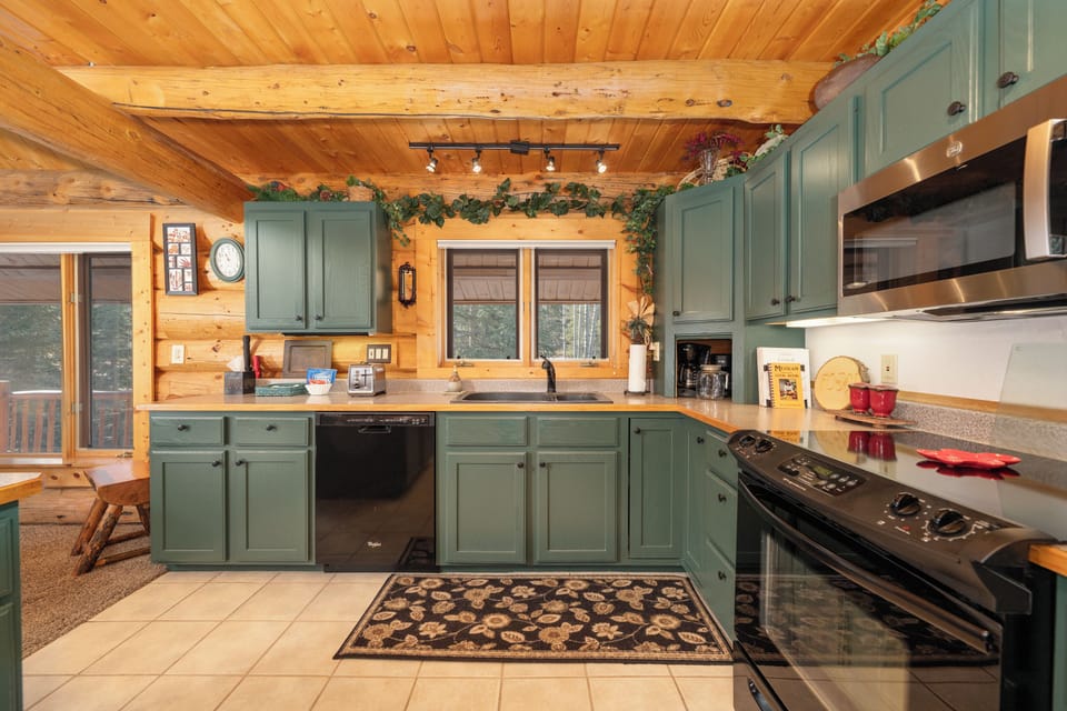 Cook memorable meals in this charming cabin kitchen with sage-green cabinets, granite counters, and rustic wood beams overhead.