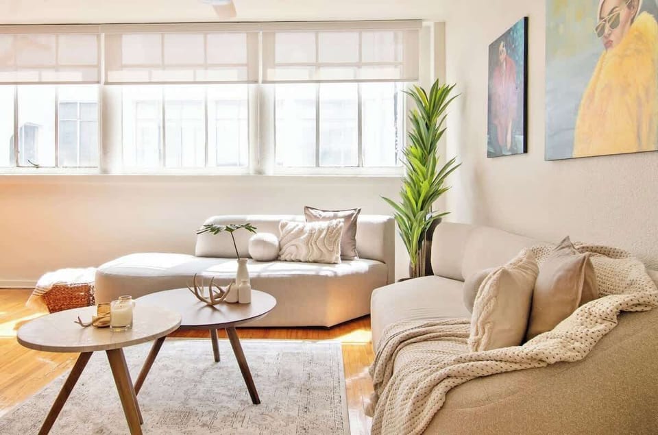 Sunlit lounge with soft neutral sofas, textured cushions, and a cozy throw. A round table with candles and greenery by the window creates a calm and inviting atmosphere beneath colorful wall art.