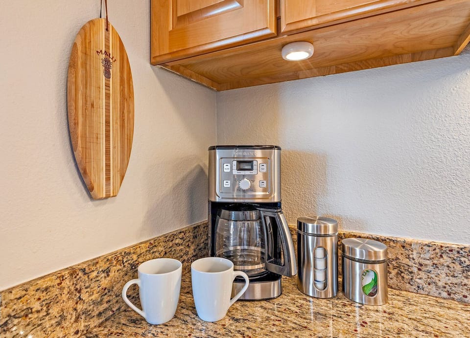 Coffee station with a drip brewer, grinder, mugs, and stainless accessories on granite counters.