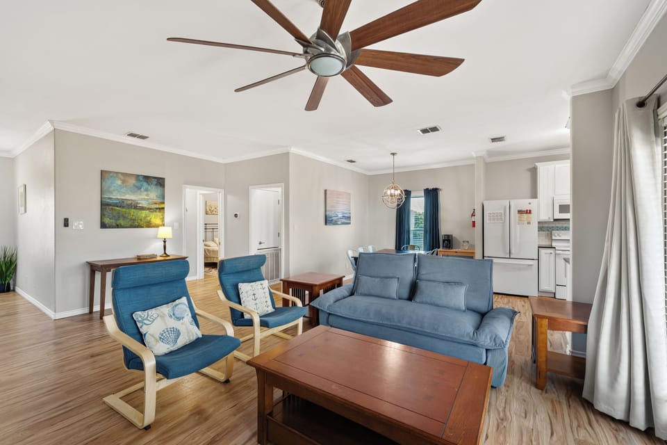 Step into your bright, open living space where coastal blues meet warm wood tones. Relax under the sweeping ceiling fan, sink into the plush sofa, and enjoy the seamless flow into the kitchen—your perfect home base for coastal escapes.