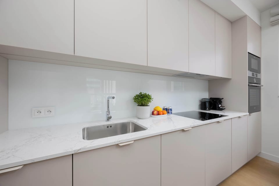 A modern white kitchen with smooth cabinets and built-in appliances. A round dining table with gray chairs sits nearby, creating a clean and practical layout.