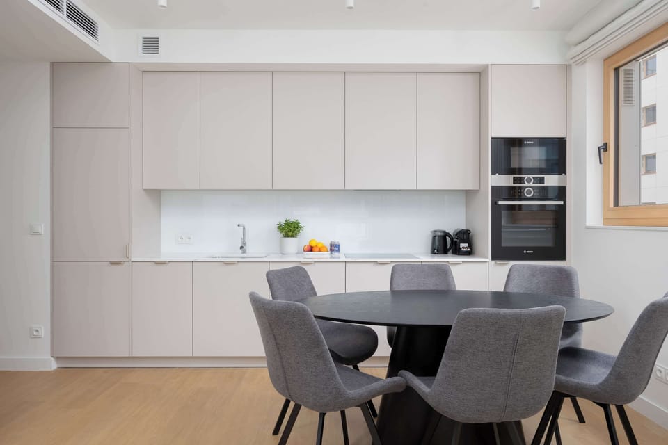 A modern white kitchen with smooth cabinets and built-in appliances. A round dining table with gray chairs sits nearby, creating a clean and practical layout.