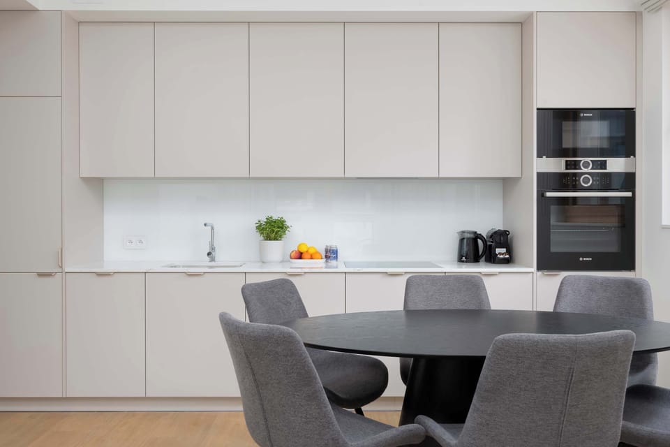 A large white kitchen wall with cabinets, a built-in oven, and a small shelf holding kitchen essentials. The dining table adds functionality without clutter.