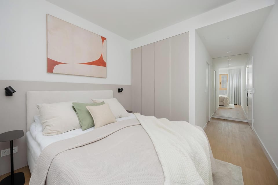 A cozy bedroom with a neatly made bed in beige tones and modern artwork above. Gentle curtains frame the window, creating a peaceful, warm atmosphere.