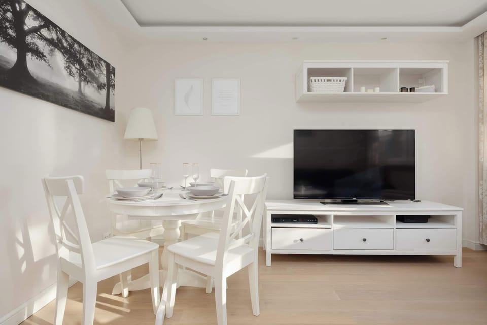 The dining area and part of the living room are captured, highlighting a small white dining table set for four. The light wooden flooring extends across the entire open space.