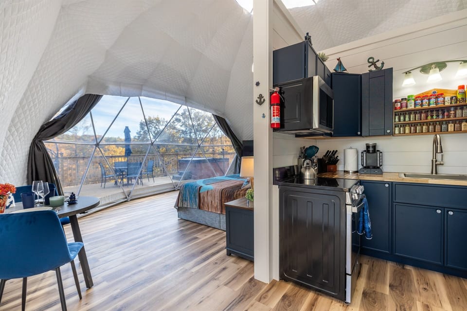 Open-layout dome with full kitchen, king bed, dining area, and floor-to-ceiling panoramic window overlooking the deck and woods