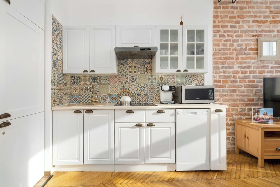 A bright kitchen featuring white cabinets, patterned backsplash tiles, and modern appliances. The space is complemented by a rustic brick wall for added character.