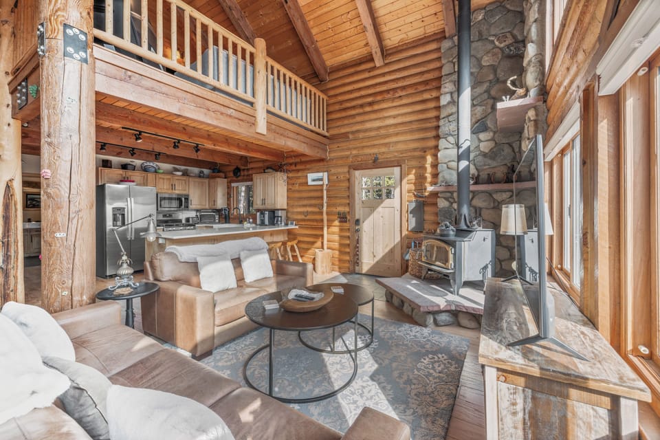 Features a wall of floor to ceiling windows, cozy leather couches centered around a Smart TV, and a wood-burning stove on a majestic stone hearth