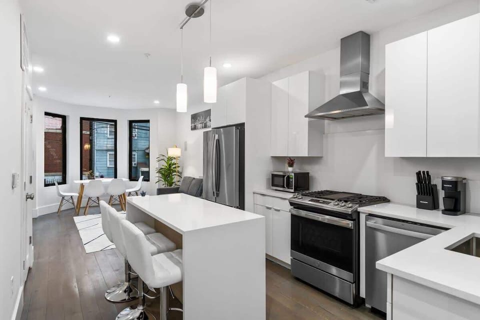 A sleek modern kitchen with glossy white cabinets, stainless steel appliances, and a large quartz island with seating for three. Clean lines, bright lighting, and a wide layout make this space both functional and stylish.