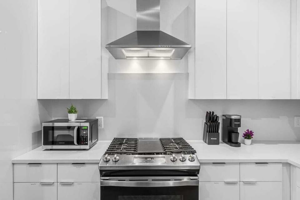 A sleek gas range sits under a stainless steel hood, framed by spotless white cabinetry. The microwave and small greenery add a simple touch while keeping the counter functional.