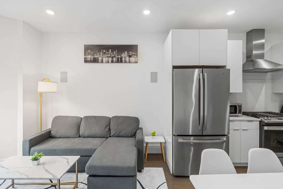 A wider shot capturing the sectional, coffee table, and stainless steel fridge just beyond. Shows the smooth flow between the living room and kitchen.