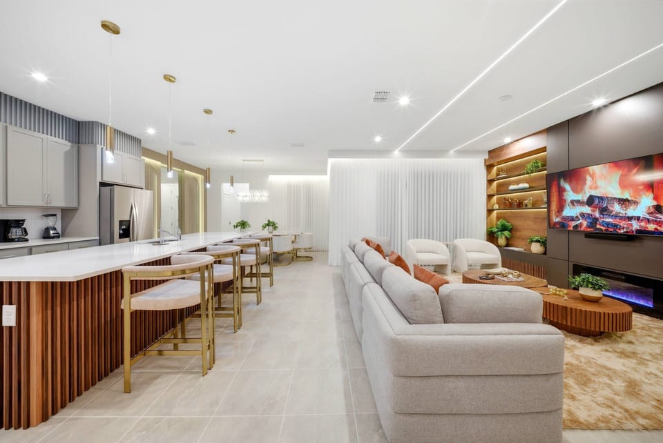 The spacious living area boasts a cozy gray sectional, a warm wood accent wall, and a modern electric fireplace, while the cushioned bar seating invites casual dining and socializing in a welcoming atmosphere.