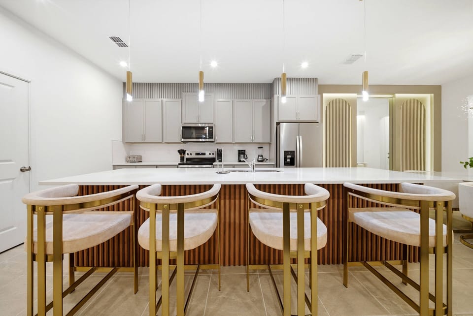 This modern kitchen features a spacious island with chic gold-accented bar stools and light wood textures, complemented by sleek cabinetry and bright pendant lights, making it the perfect setting for your culinary adventures.