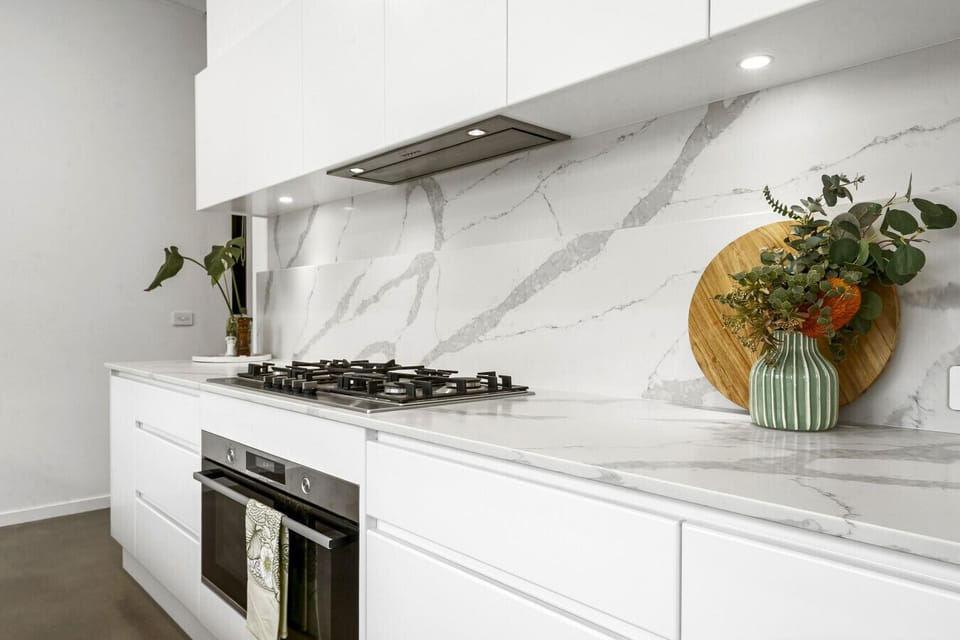 Fully equipped kitchen with a gas cooktop, oven, and rangehood set against a marble benchtop and full-height splashback. Sleek white cabinetry offers plenty of storage and prep space for cooking.