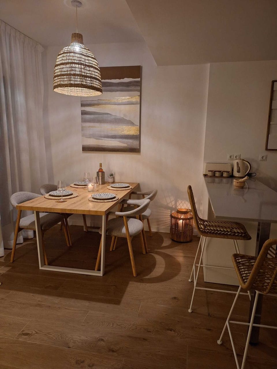 Modern dining space with a wooden table, cushioned chairs, and soft pendant lighting—framed by abstract art, wicker bar stools, and cozy, earthy vibe perfect for casual meals and relaxed conversations.