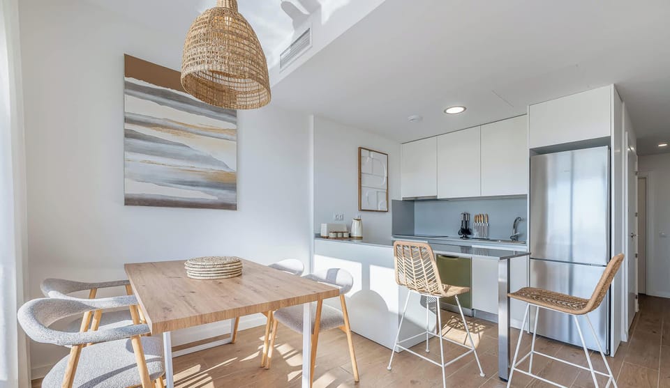 This bright kitchen and dining area features sleek, white cabinetry and stainless steel appliances. The dining space has a modern wooden table, and the design is accented by a woven light fixture and counter seating.