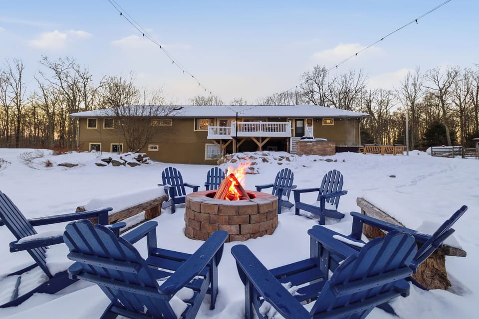 Snowy backyard fire pit with Adirondack seating and a cozy home view.