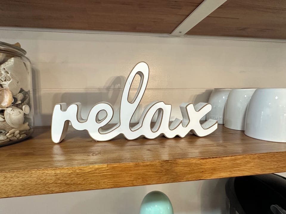 Wood shelf with seashell jar, mugs, and “relax” sign in beachy neutrals.