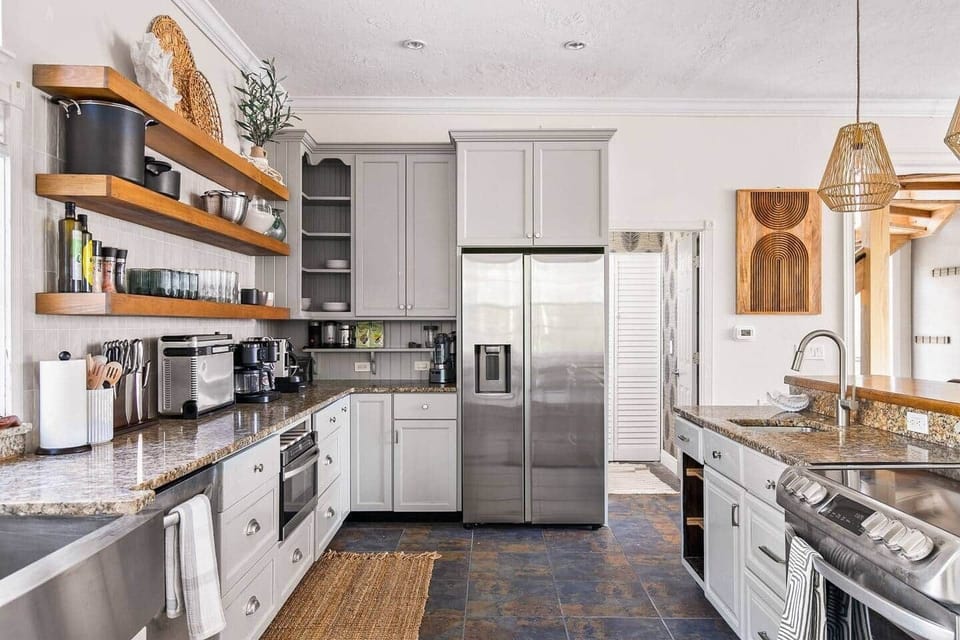 Chef's kitchen with stainless appliances, granite counters, gray shaker cabinets, floating wood shelves, and fully stocked cookware.