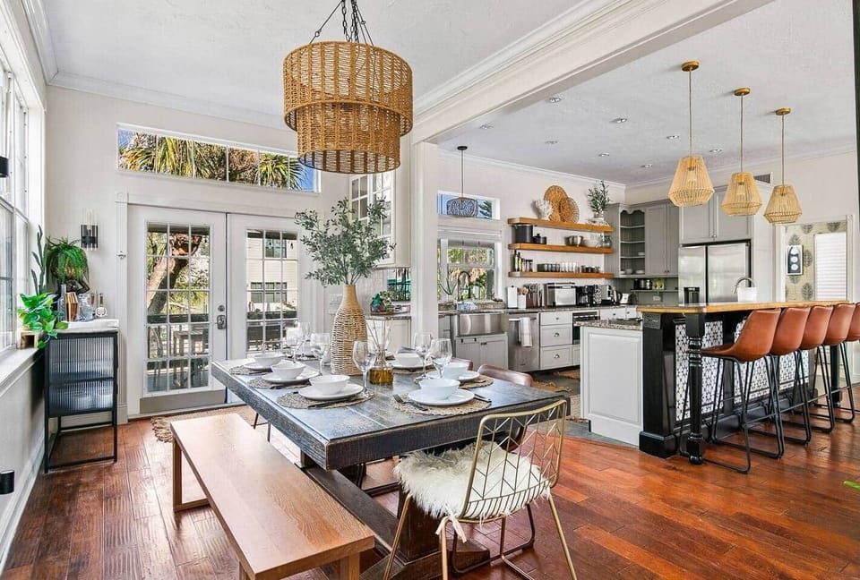Open-concept dining with farmhouse table, wicker chandelier, French doors to deck, and seamless flow into the chef's kitchen.