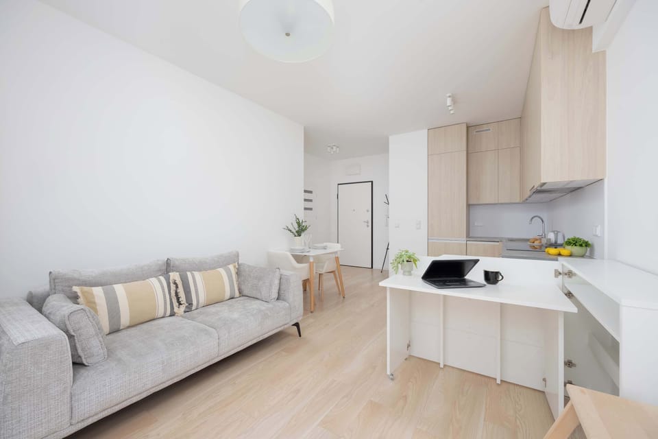 A bright living area with a gray sofa and a small dining table. The space includes a kitchenette with light wood cabinets and modern appliances.