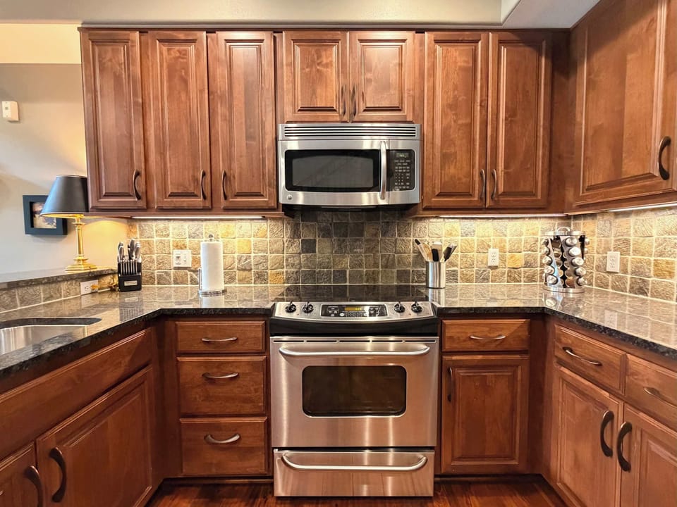 Kitchen with stainless range, stone tile backsplash, dark granite counters, and rich wood cabinetry — fully equipped for real cooking