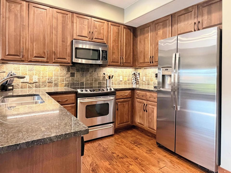Bright full kitchen with stainless fridge, granite counters, stone tile backsplash, and hardwood floors — more counter space than you'd expect