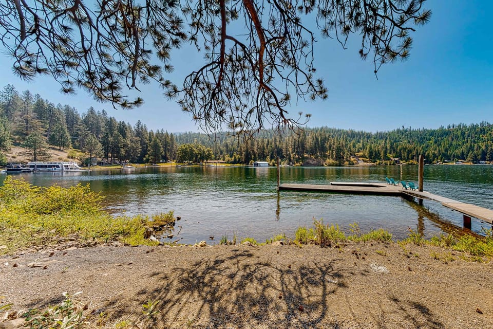 Bright lakeside dock with Adirondack chairs, pine-tree framing, and panoramic water and mountain views