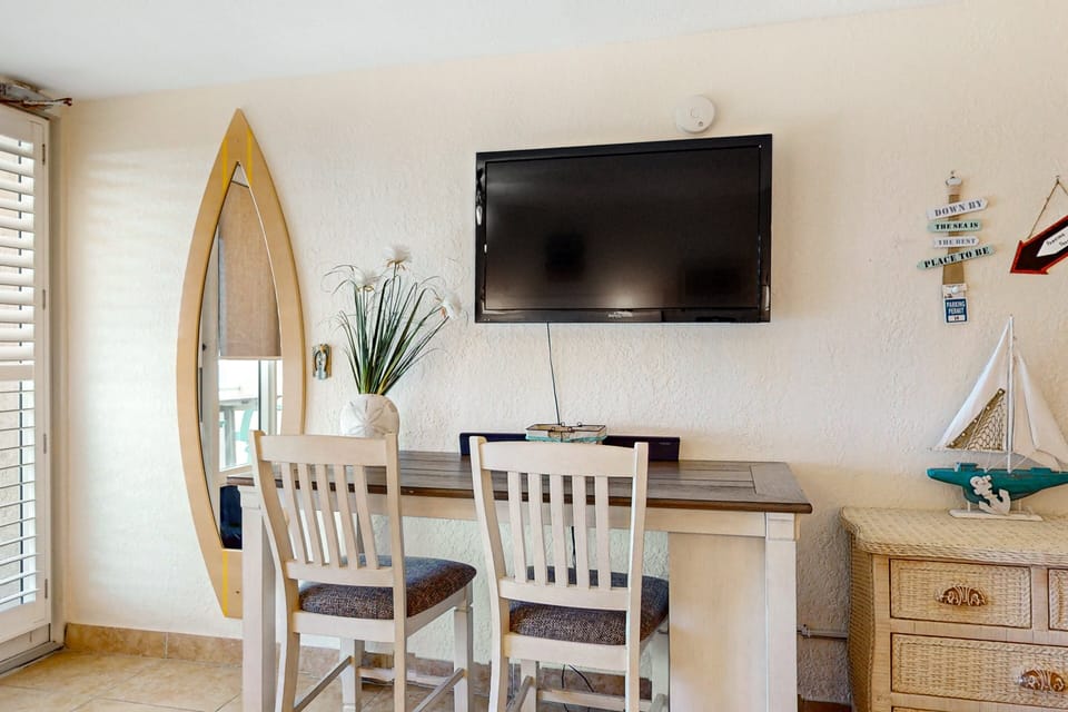 Cozy dining nook with coastal décor, seating for two, and wall-mounted TV. Perfect for a relaxed beach stay, offering a charming spot to dine, unwind, and enjoy your favorite shows after the beach.