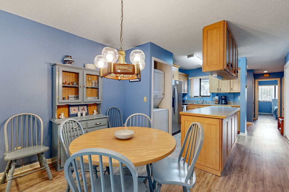 Charming beach-style dining space with round table seating and open access to a fully equipped kitchen—perfect for family meals and gathering after a day at the shore during your relaxing beach stay getaway.