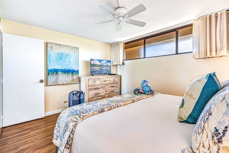 Relax in a bright bedroom featuring a comfortable bed with soft neutral linens, accented by coastal artwork and natural light streaming through wide windows.