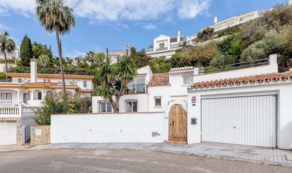 Mediterranean-style houses, terracotta roofs, palm trees, and lush greenery. A curved street passes a home with an arched wooden door and garage, creating a warm, inviting scene.