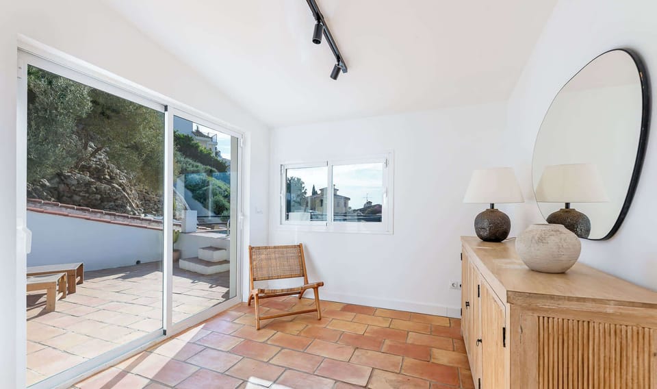 Bright room with terracotta floors, sliding glass doors to a patio, rustic sideboard with lamps and mirror, and a woven chair. Minimalist design and natural light create a warm, airy atmosphere.