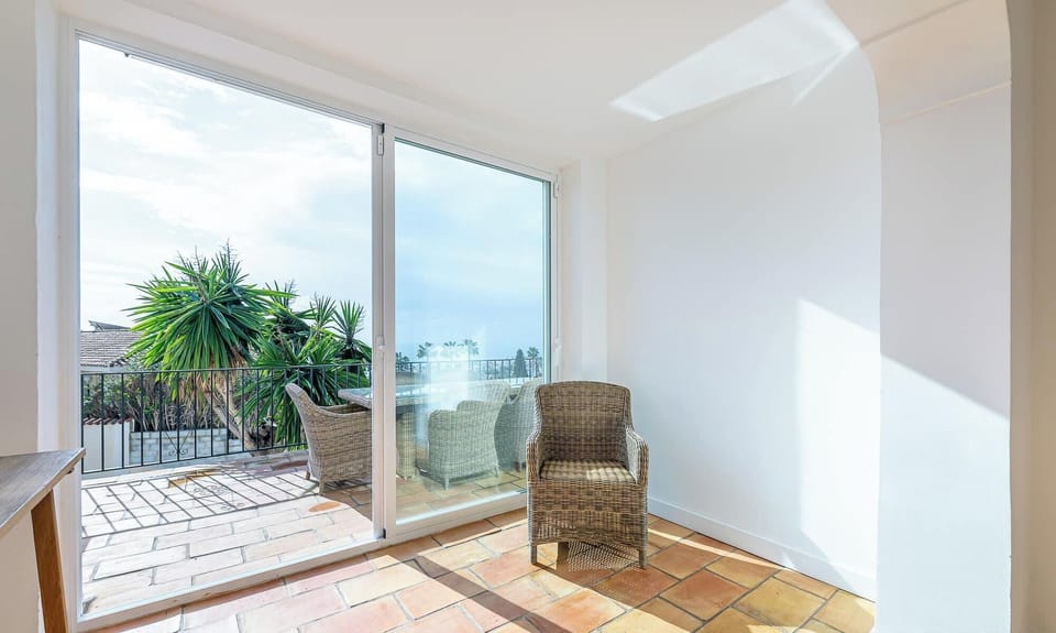 This airy space features terracotta floors, a glass sliding door opening to a lush patio with wicker furniture, and sunlight streaming in. A wicker chair inside faces the greenery, creating a bright, relaxing atmosphere perfect for unwinding. 