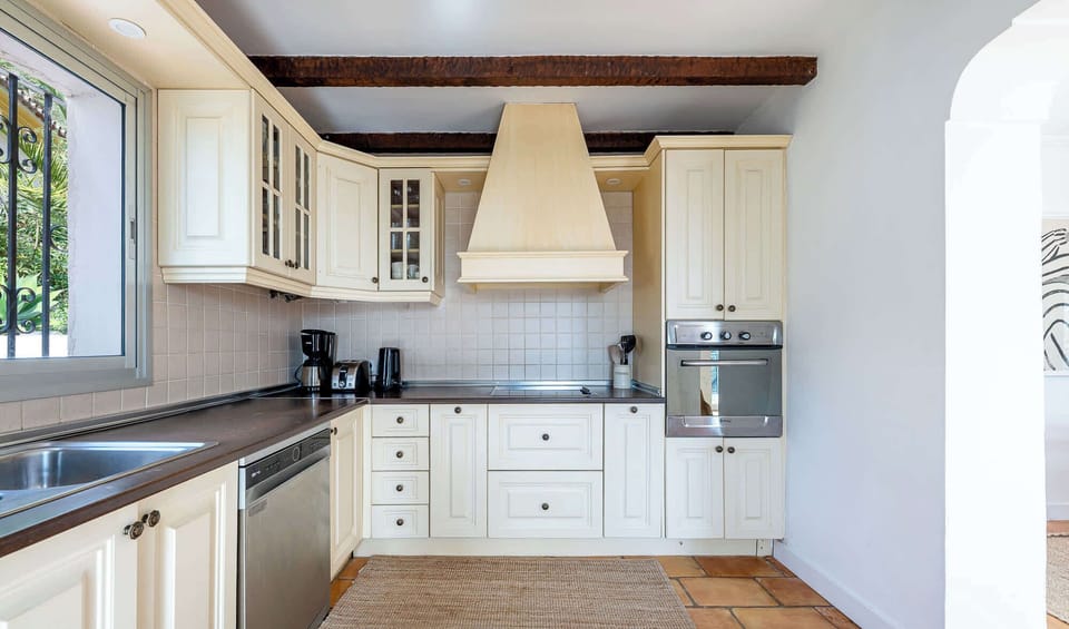 Modern kitchen with cream cabinets, dark counters, stainless appliances, terracotta floors, and wooden beams. Warm rustic touches blend with sleek design, creating a stylish and inviting cooking space.