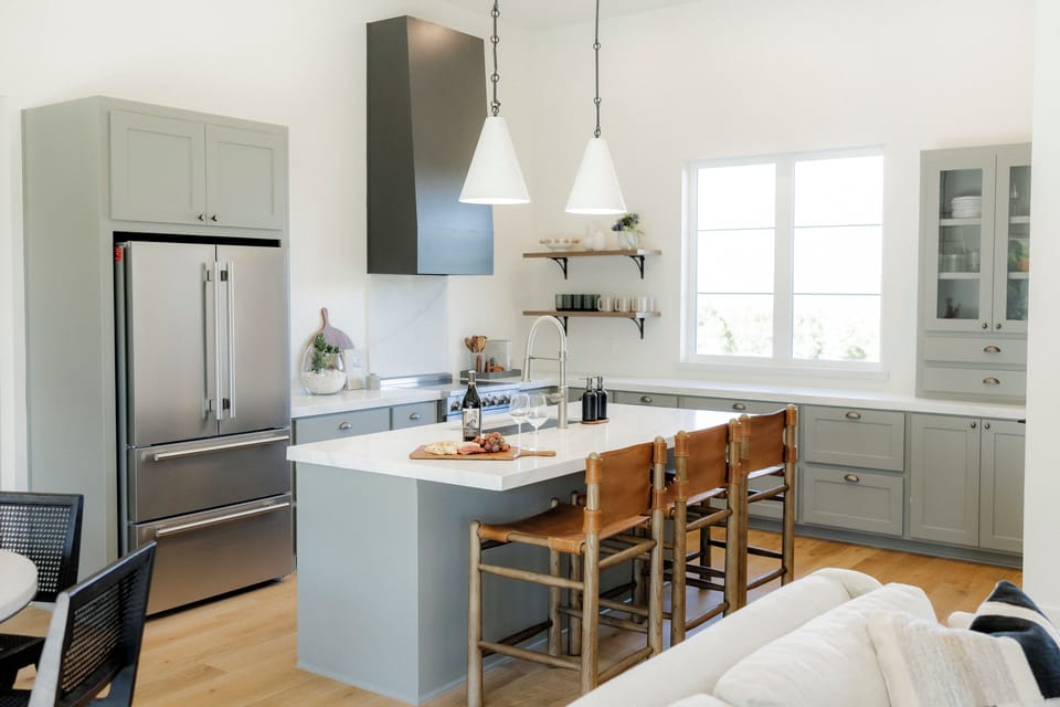 Airy chef’s kitchen with island seating, stainless fridge, and open shelving.
