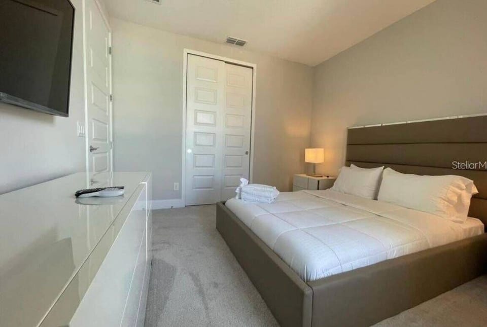 Experience a cozy bedroom featuring soft, neutral tones and plush bedding that welcomes relaxation. Modern furnishings, like a stylish bedside lamp and wall-mounted TV, enhance the comfortable atmosphere ideal for unwinding.