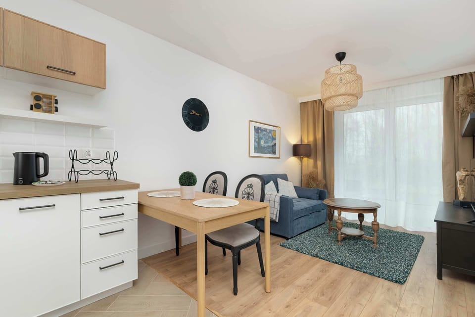 A bright dining and living area featuring a small wooden dining table with chairs and a cozy sofa. Natural light fills the space through large windows.