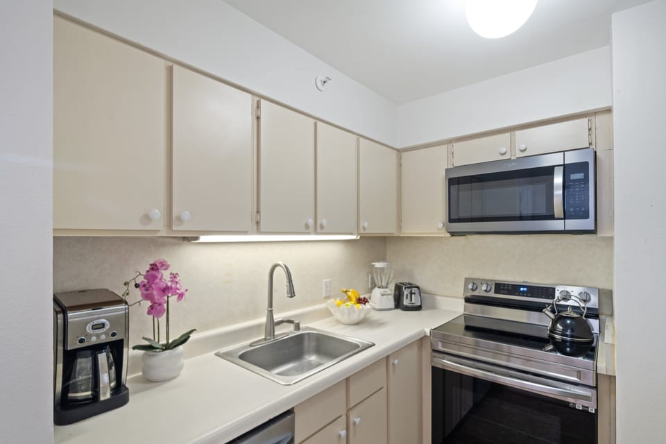 Compact kitchen with beige cabinetry, granite counters, and modern appliances accented by a potted orchid.