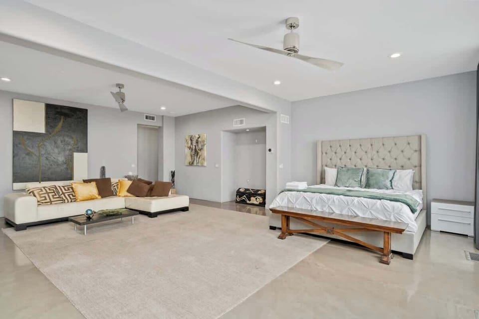 This beautifully designed bedroom features a neutral-colored sectional sofa and a king-sized bed with a tufted headboard for a touch of elegance. Wide windows with sheer curtains allow for soft light, while blackout curtains cater to day sleepers.