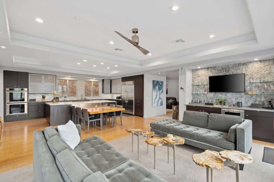This tastefully designed open-concept main area features wood-finished flooring, white walls, and ceiling. Pin lights highlight the accent wall, while tufted gray sofas, a wooden centerpiece, and top-of-the-line kitchen appliances complete the space