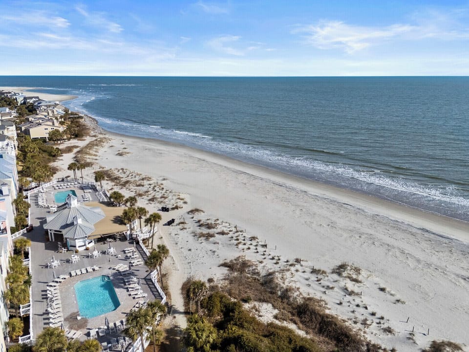 A peaceful stretch of sandy shoreline lined with coastal homes and a sparkling pool overlooking the endless blue horizon.