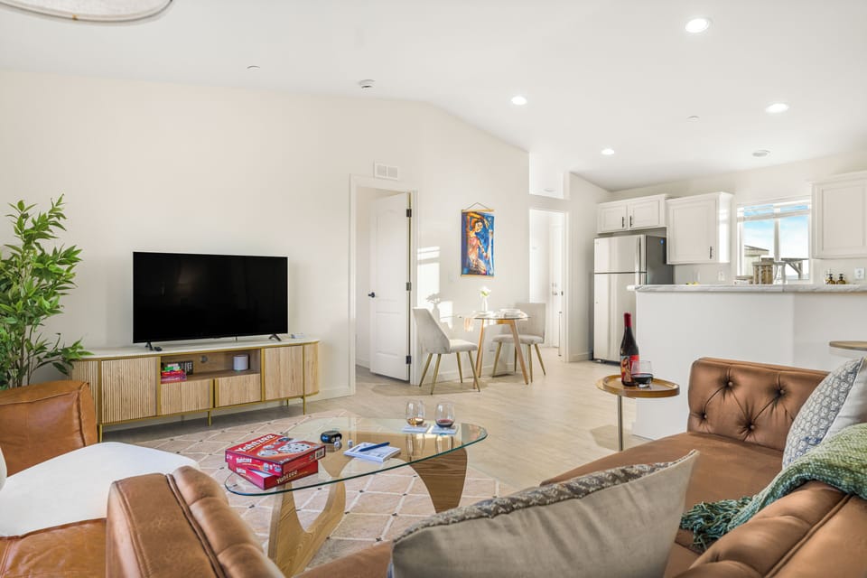 Open-plan living room with white walls, a mounted TV, and seating leading into the kitchen.