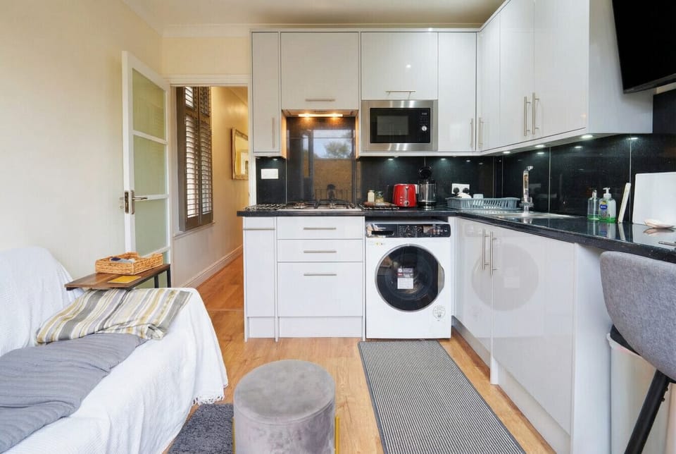 The open-plan kitchen is fitted with gloss white units, dark worktops, a gas hob, integrated microwave, and a washing machine, with a sofa visible to the left.