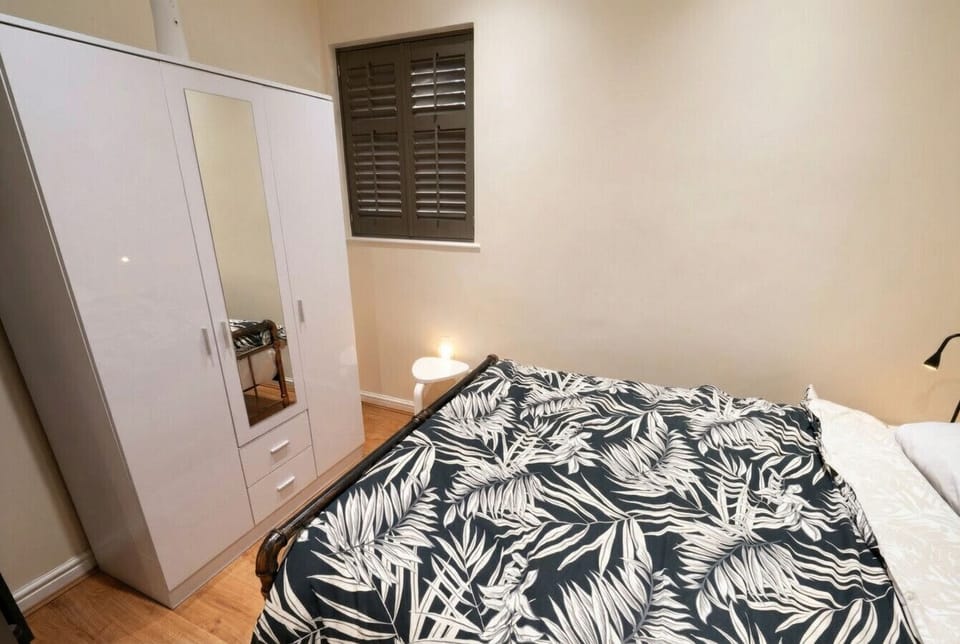 The bedroom includes a full-height white wardrobe with a mirrored door, a bedside lamp, and a shuttered window providing privacy.