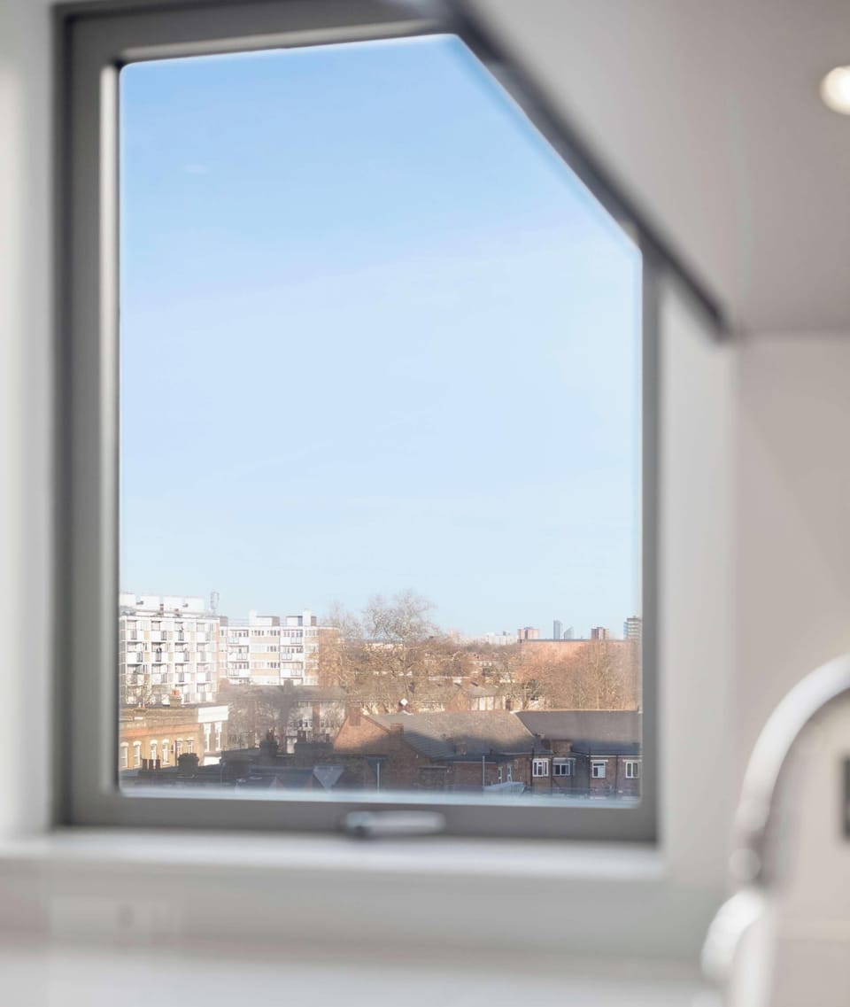 A modern kitchen sink area features a sleek chrome faucet and white countertop. A large, uniquely shaped window overlooks a cityscape of brick buildings under a clear blue sky.