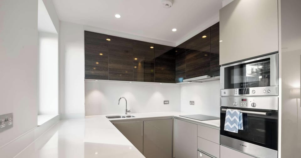 This modern U-shaped kitchen features a sleek, minimalist design with glossy white countertops and two-toned cabinetry. Dark wood accents and stainless steel appliances create a clean, sophisticated aesthetic.